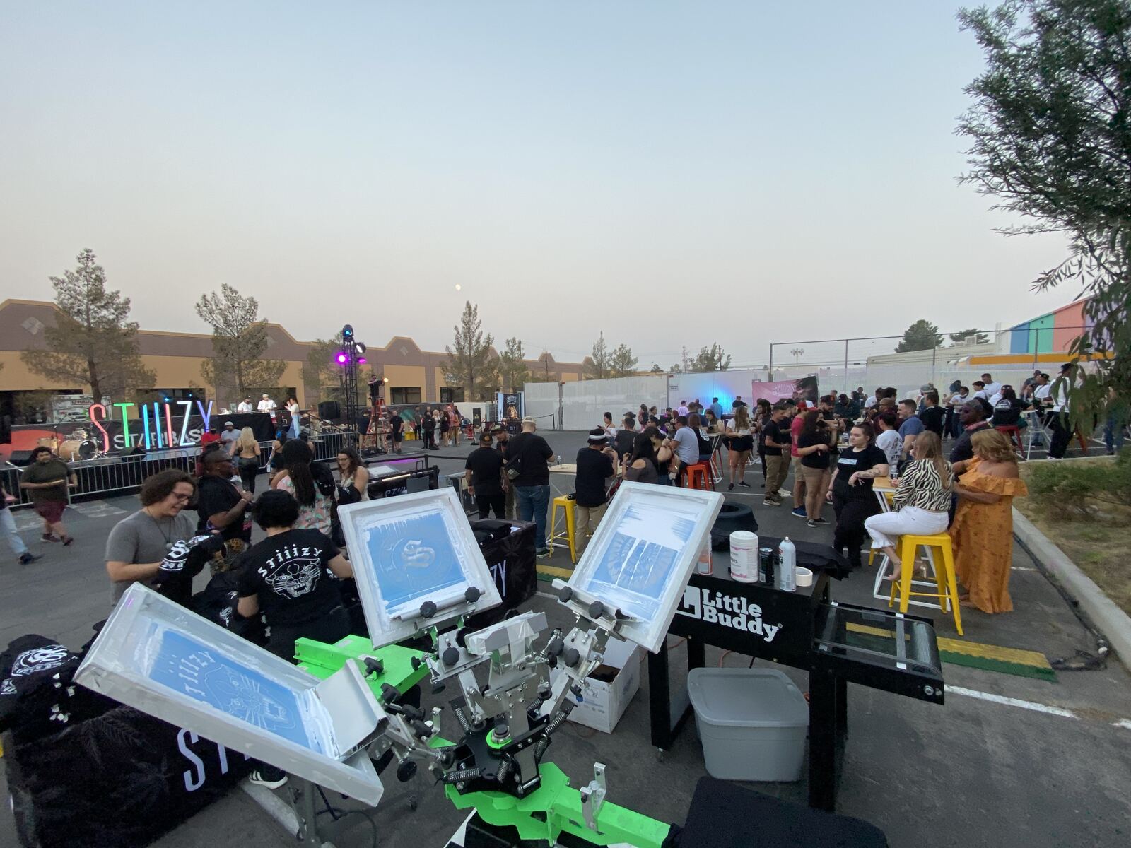 Outdoor festival scene with a live printing equipment demonstration, numerous attendees, and branded signage